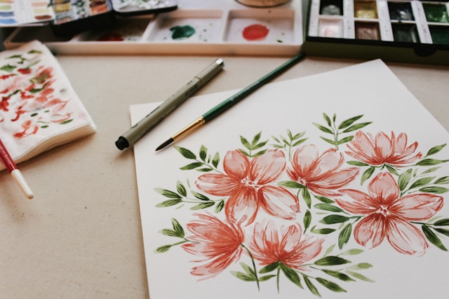 Watercolor painting of pink flowers on white paper, with art supplies nearby, by Mia, age 30.